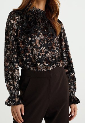 Schwarze durchscheinende Bluse mit braunem und beigem Blumen-Paisley-Muster, hohem Rüschenkragen und langen Puffärmeln, kombiniert mit dunkler Hose.