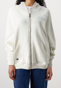 Felpa bianca con zip e finitura testurizzata, dotata di tasca frontale, dettagli in argento e logo "PEQUS" in azzurro chiaro sul petto.