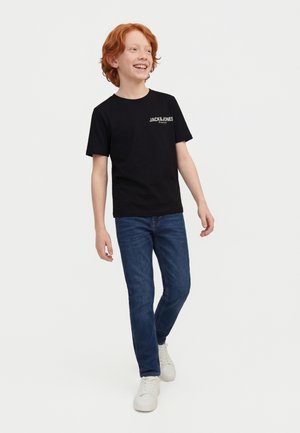 Ragazzo dai capelli rossi che cammina, sorridendo, indossando una T-shirt nera Jack & Jones, jeans blu e sneakers bianche su sfondo chiaro e uniforme.