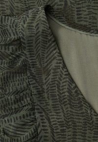 Tissu vert olive foncé avec des motifs linéaires noirs, montrant un gros plan sur l'encolure cousue et des sections pliées d'un vêtement texturé.
