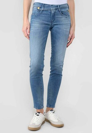 Jean en denim bleu taille haute, coupe slim, avec détail de boutons dorés, finition délavée et deux poches avant, associés à des baskets blanches.