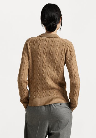 Cable-knit sweater in taupe with ribbed hem and cuffs. Worn with grey trousers, showcasing textured yarn and fitted design.