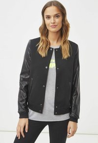 Chaqueta bomber negra con un cuerpo de tela texturizada y mangas de cuero negro brillante. Cuenta con botones de presión y un dobladillo acanalado. Llevada sobre una camiseta gris.