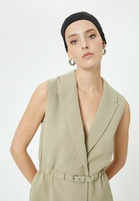 Helles olivgrünes ärmelloses Jumpsuit mit tiefem V-Ausschnitt, tailliertem Gürtel und strukturierten Revers. Glattes Material mit lässiger Passform.