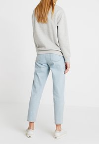 Woman with long hair wearing a grey sweatshirt, light blue high-waisted jeans, and white platform sneakers standing facing away.