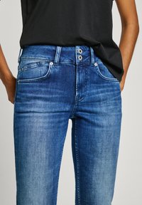 Jean en denim de lavage bleu moyen à foncé. Dispose de cinq poches, de deux fermetures à bouton et d'une légère décoloration au niveau des cuisses.