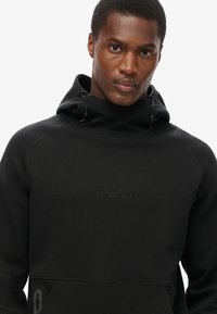 Sudadera negra con cuello alto, logo sutil y bolsillo con cremallera. Hecha de una suave tela texturizada con cordones en la capucha.