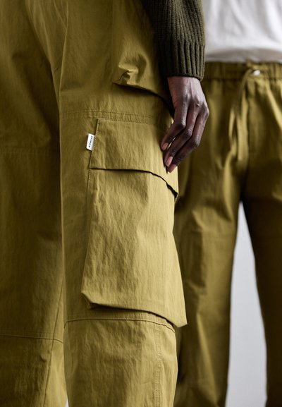 Olive green cargo pants with multiple pockets, soft texture, and a relaxed fit; features a visible brand label on the side seam.