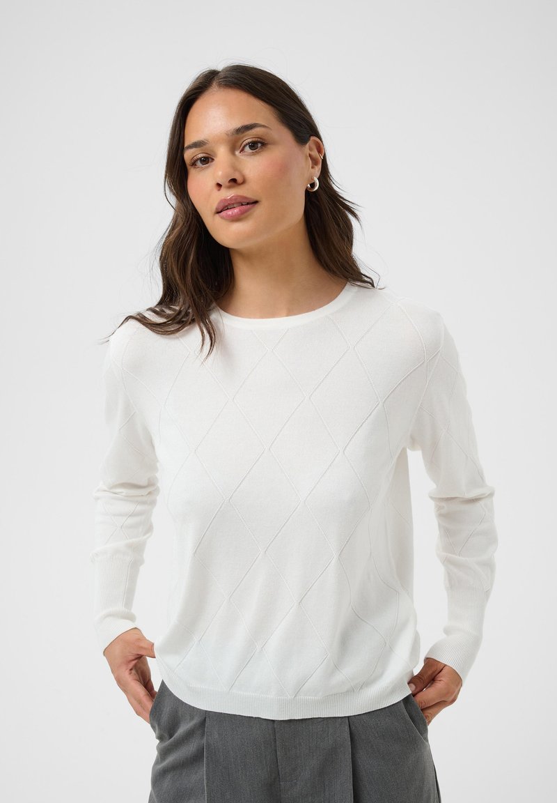 Femme portant un pull blanc à motif losange et un pantalon gris, les mains dans les poches, debout devant un fond clair uni.