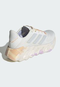 Scarpa sportiva bianca con tomaia in mesh testurizzato, accenti grigi e suola intermedia a gradiente colorato. Presenta tre strisce riflettenti e supporto morbido per il tallone.