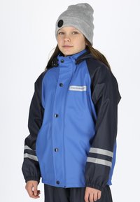 Blå vandtæt jakke med mørkeblå ærmer, sølvstriber ved manchetterne, trykknaplukninger og et logo patch. Sammen med en grå strikket beanie.