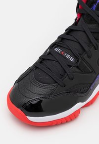 Czarny but sportowy z teksturowaną skórzaną cholewką, błyszczącymi akcentami z lakierowanej skóry, czerwoną podeszwą oraz kolorowymi oczkami z czarnymi sznurówkami.