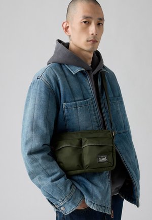Man wearing a blue denim jacket over a gray hoodie, carrying an olive green crossbody bag with front pockets and a logo patch.