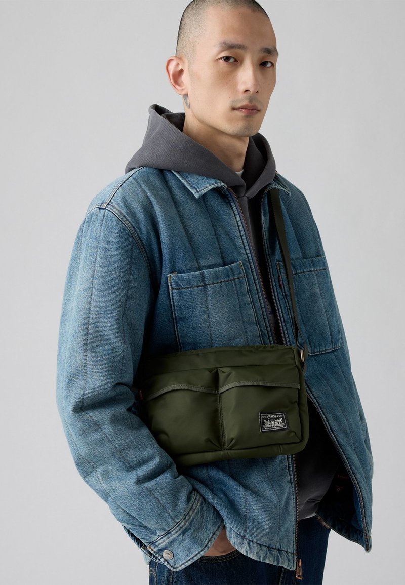 Uomo che indossa una giacca di jeans blu sopra un hoodie grigio, con una borsa a tracolla verde oliva con tasche frontali e una patch con il logo.