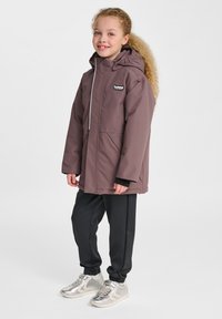 Lila wasserdichte Jacke mit Kapuze, zwei Fronttaschen und einem Logo-Patch. Kombiniert mit schwarzen Jogginghosen und silbernen Sneakers.