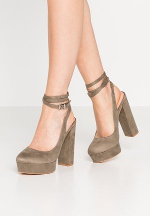 Nogi w taupe zamszowych platformach na wysokim obcasie z paskami wokół kostki i klockowymi obcasami na białej podłodze i tle.