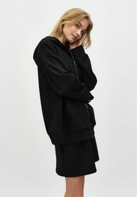 Svart hoodie med dragkedja, oversize passform, mjukt material och sidofickor, matchad med svarta shorts. Enkelt och minimalistiskt design.