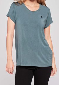 Tyrkysové tričko s krátkým rukávem a kulatým výstřihem, s jemnou texturou a vyšívaným logem na levé straně hrudi. Kombinované s černými kalhotami.