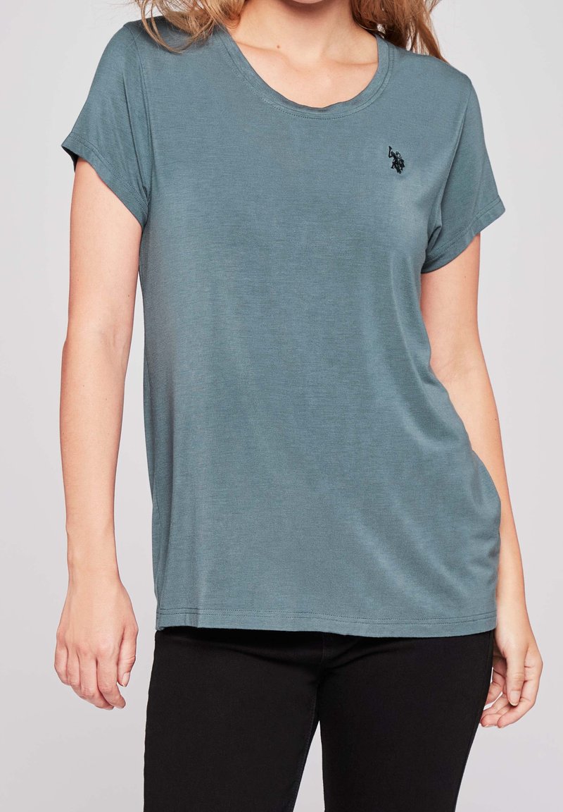 Tyrkysové tričko s krátkým rukávem a kulatým výstřihem, s jemnou texturou a vyšívaným logem na levé straně hrudi. Kombinované s černými kalhotami.