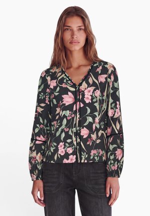 Femme aux longs cheveux portant un chemisier noir à fleurs roses et vertes et un jean foncé, debout devant un fond blanc.