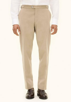 Mann trägt beige maßgeschneiderte Hosen, weiße Hemdärmel und dunkelbraune Lederschuhe stehend.