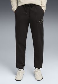 Sorte jogger-bukser med snøre i taljen, sidelommer og cremefarvet logo-detalje på låret; parret med lyse sneakers.