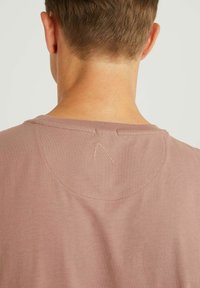 Světle růžové tričko s jemnou texturou, které má kulatý výstřih a malý vyšitý trojúhelníkový detail na zadní straně.