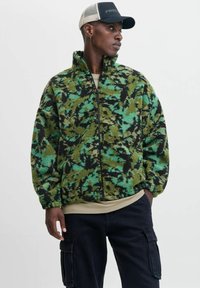 Groene en zwarte fleecejack met een hoge kraag, ritssluiting aan de voorkant en een camouflagepatroon. Draag het over een beige shirt en combineer het met donkere broeken.