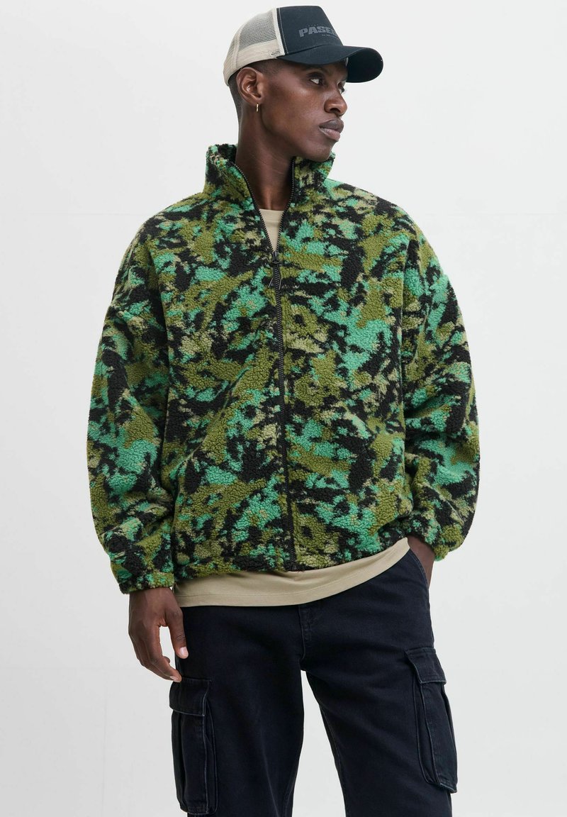 Groene en zwarte fleecejack met een hoge kraag, ritssluiting aan de voorkant en een camouflagepatroon. Draag het over een beige shirt en combineer het met donkere broeken.