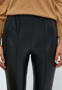 Leggings neri in ecopelle con una texture liscia e cuciture verticali, abbinati a un maglione marrone chiaro. I dettagli includono un design a vita alta.