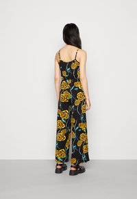 Macacão preto de perna larga com padrão floral amarelo, alças finas ajustáveis e um corte descontraído. Tecido texturizado com acabamento suave.
