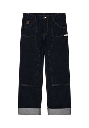Persoon die donkere spijkerbroek draagt met opgerolde pijpen, bruine werklaarzen, een wit overhemd en een donker spijkerjasje.