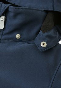 Nahaufnahme des Kragens einer blauen Jacke mit einem Druckknopf und Nähdetails, die die Textur des Stoffes und Designelemente zur Geltung bringen.