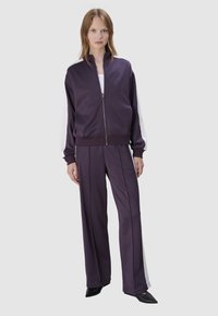 Paarse sportieve zip-up jas met witte accenten, gecombineerd met bijpassende paarse wijde broek. Gemaakt van glad, ademend materiaal.