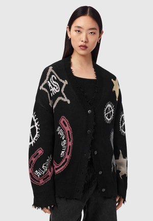 Kvinne med rett mørkt hår iført svart knappet cardigan med beige stjerner, hvite fredssymboler og rosa hestesko på ermene og fronten.