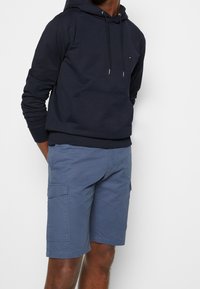 Marineblaue Kapuzenpullover mit Kordelzügen, Fronttasche und gerippten Bündchen, kombiniert mit hellblauen Cargo-Shorts mit Seitentaschen.