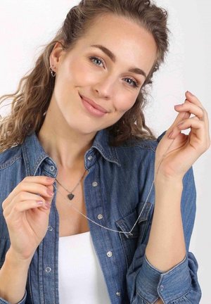 Zilveren kettingen met een hartbedel, gelaagd ontwerp, en een model dat een denim overhemd draagt, die de details en glans van de sieraden toont.