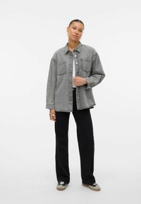 Giacca di denim grigia con due tasche sul petto, chiusura a bottoni e una vestibilità rilassata. Abbinata a pantaloni neri e sneaker grigie. Sfondo semplice.