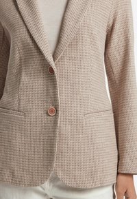 Vävd kavaj i en ljusbrun nyans, med ett texturerat mönster, slagkragar, två fickor och en tvåknapps stängning.