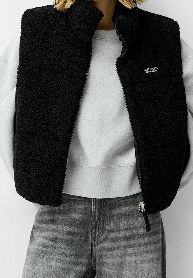 Zwarte fleece bodywarmer met hoge kraag, ritssluiting en gestructureerde stof. Draagt over een lichtgrijze sweatshirt en gecombineerd met grijze spijkerbroek.