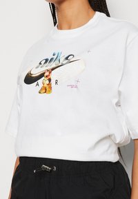 Person i vit Nike Air t-shirt med en flerfärgad gelébjörn-grafik och svarta byxor med dragsko i midjan.