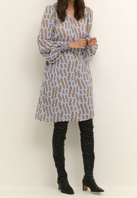 Šaty s potiskem, které mají výstřih do V a dlouhé rukávy s volány, v hnědých a světle modrých geometrických tvarech, doplněné nadkolenkovými černými botami.