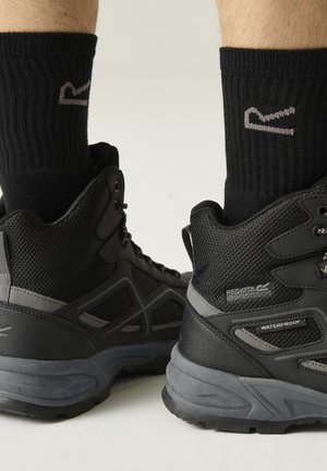 Zwarte waterdichte wandelschoenen met grijze zolen, gedragen met zwarte crewkousen op een lichte achtergrond.