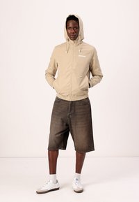 Beige lättviktsjacka med huva och framzipper, i kombination med mörka denimshorts och vita sneakers. Enkel logotypdetalj på jackan.