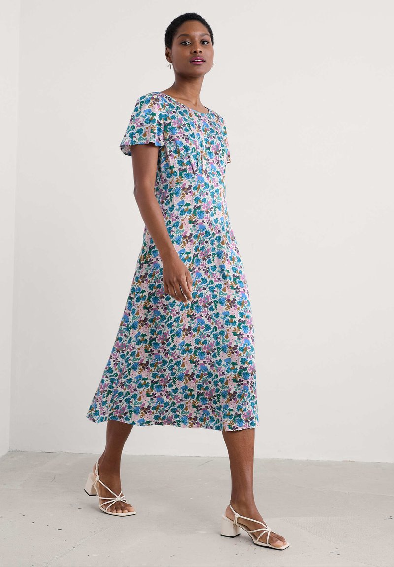 Robe maxi florale en tissu léger avec des manches courtes à volants, des motifs violets, bleus et verts, et une coupe décontractée ; portée avec des sandales à talons carrés.