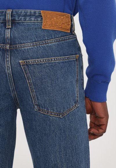 Blue denim jeans feature a brown leather label at the waistband, contrasting stitching, and a back pocket with a distinct shape.