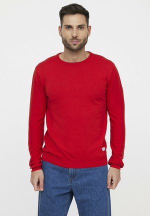 Pull encolure ronde rouge en tissu doux, avec des manches longues et un ourlet côtelé. Porté avec un jean en denim bleu.