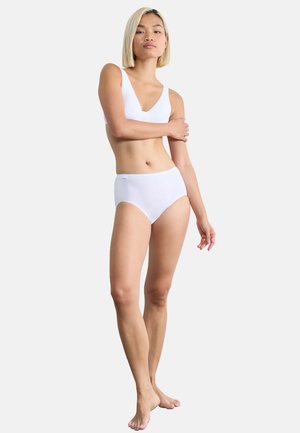 Soutien-gorge sans fil blanc avec un décolleté en V profond et culottes taille haute, fabriqués en tissu doux. Le mannequin est pieds nus, les bras croisés. Texture lisse.
