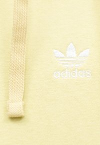 Žltá bavlnená mikina so zapusteným bielym výšivkovým logom Adidas a textúrovaným šnúrkou. Detailný záber na detaily látky a šitia.
