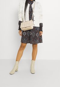 Valkoinen neulottu neuletakki tumman paisley-mekon päällä, jossa on röyhelösateet, yhdistettynä vaaleisiin nilkkasaappaisiin ja vaaleanbeigeen olkalaukkuun.